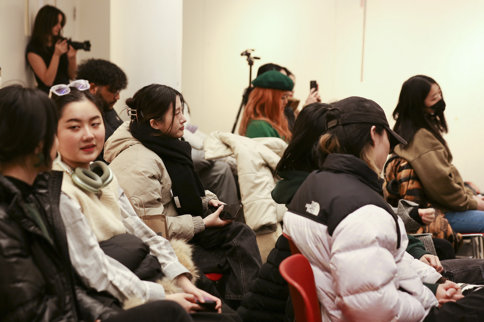 Seated students are in a room, some appear to be talking to another another, some do not appear to be talking while a person holds a camera near the edge of the frame